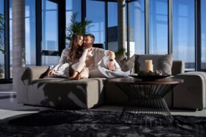 Front view of happy couple lying on sofa and hugging near large panoramic window with incredible view on city. Concept of enjoying free time at home together.
